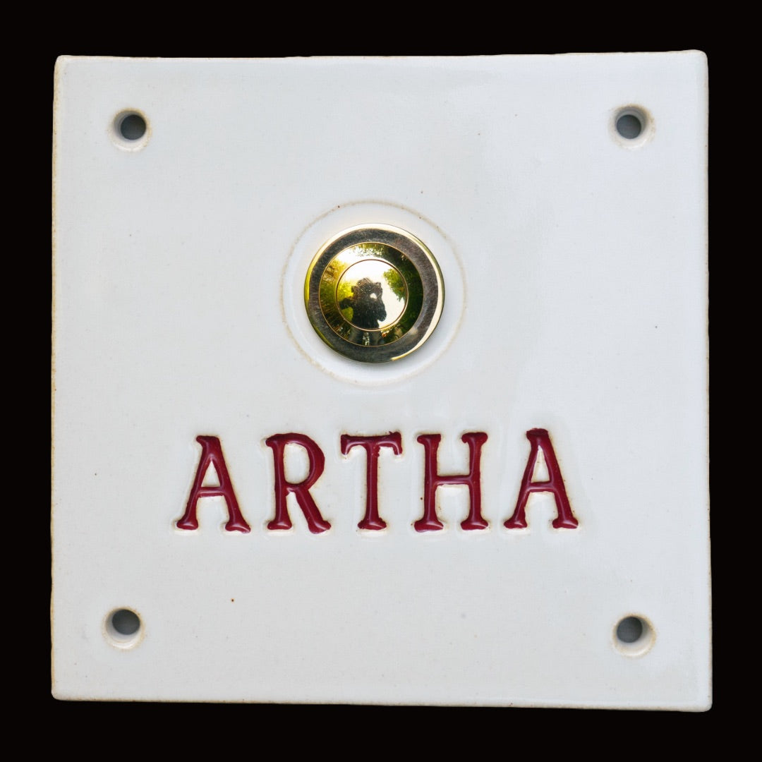 Handgefertigte Keramik-Türklingel in Weiß mit goldenem Klingelknopf. Der Name Artha ist in tiefroter Schrift eingraviert und verleiht Ihrem Eingangsbereich zeitlosen, persönlichen Charme