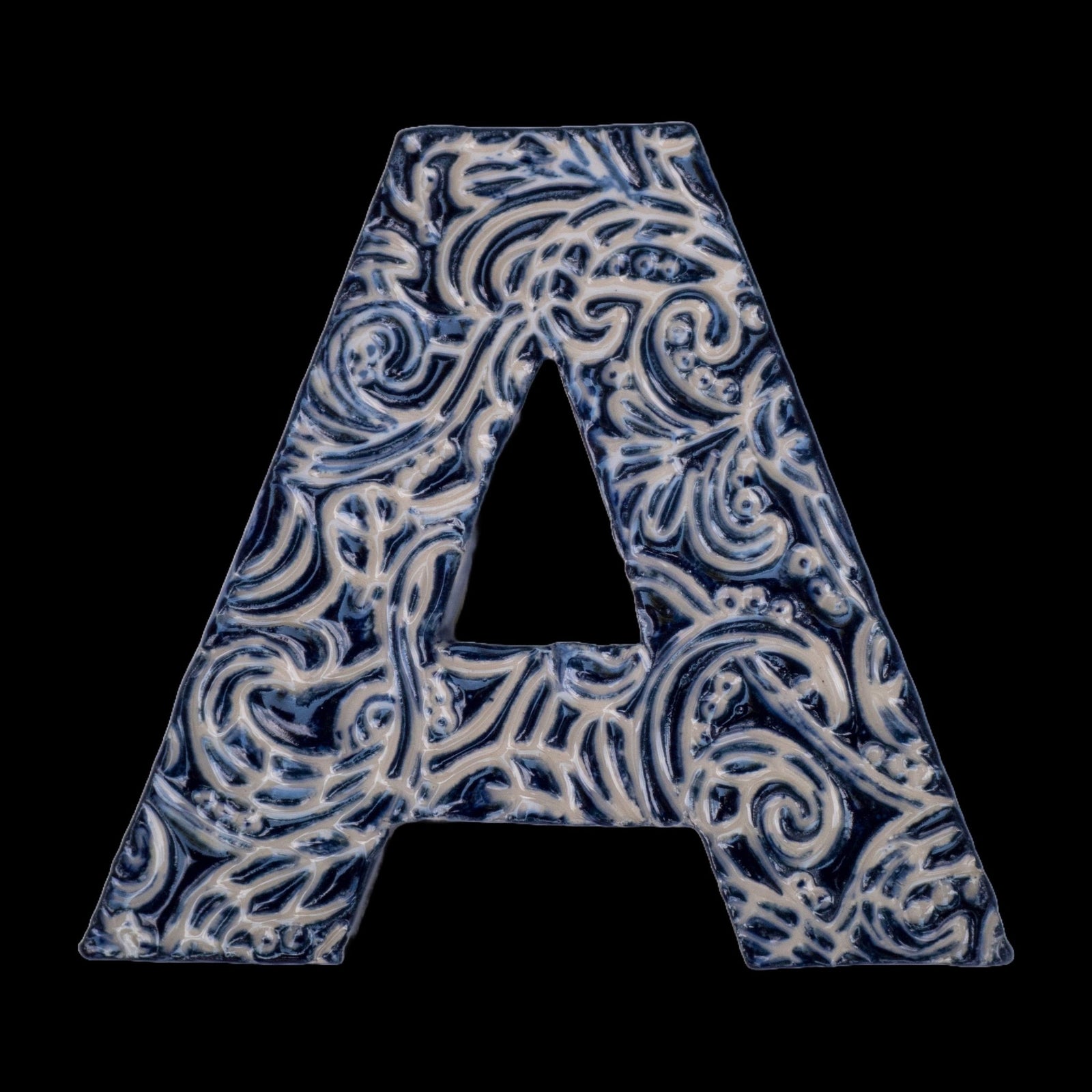 Handgefertigter Keramikbuchstabe „A“ in Dunkelblau mit beigefarbenen Ornamentmustern, vor schwarzem Hintergrund dargestellt.