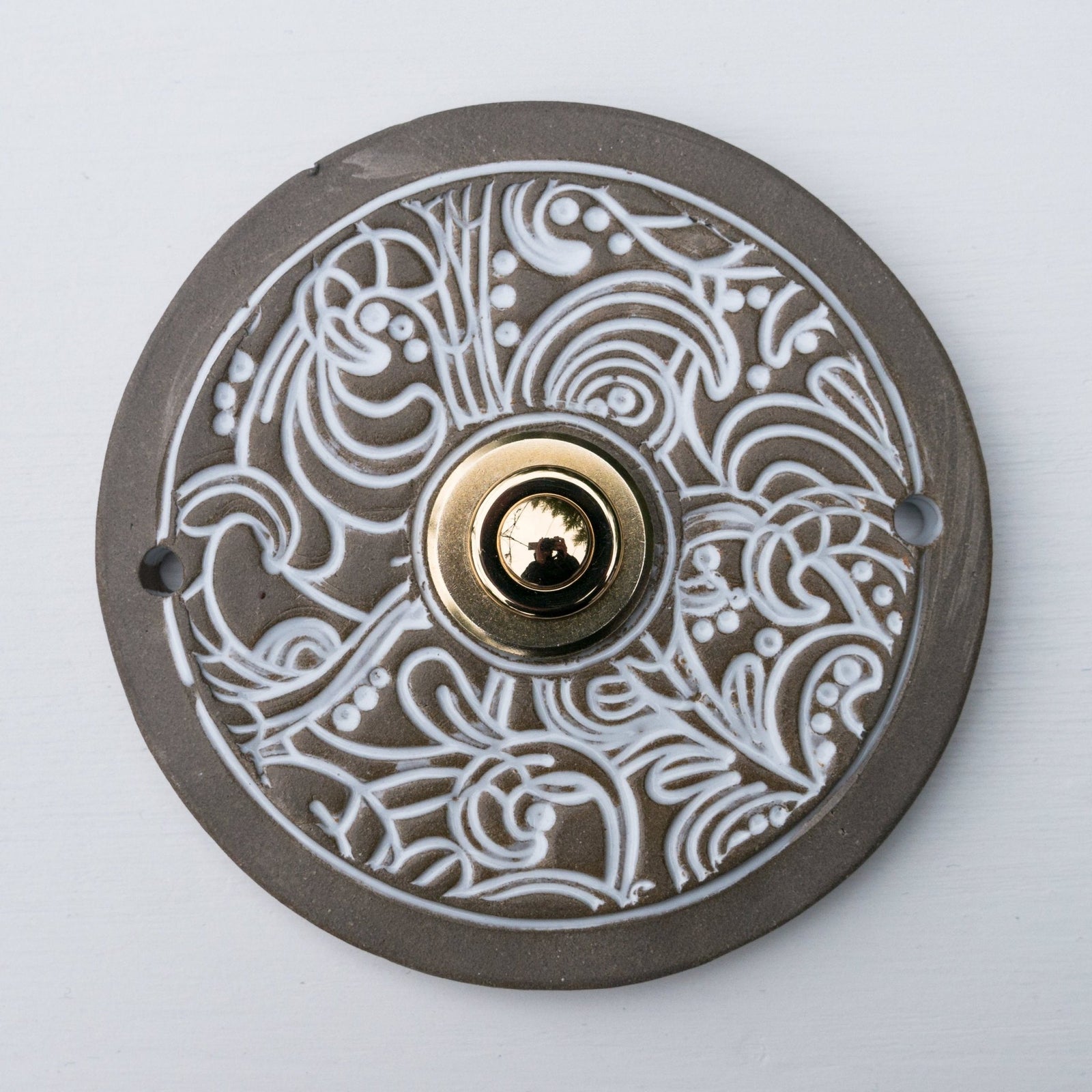 Runde dekorative Klingel mit filigranem weißen Wirbelmuster auf grauer Platte und goldenem Druckknopf in der Mitte