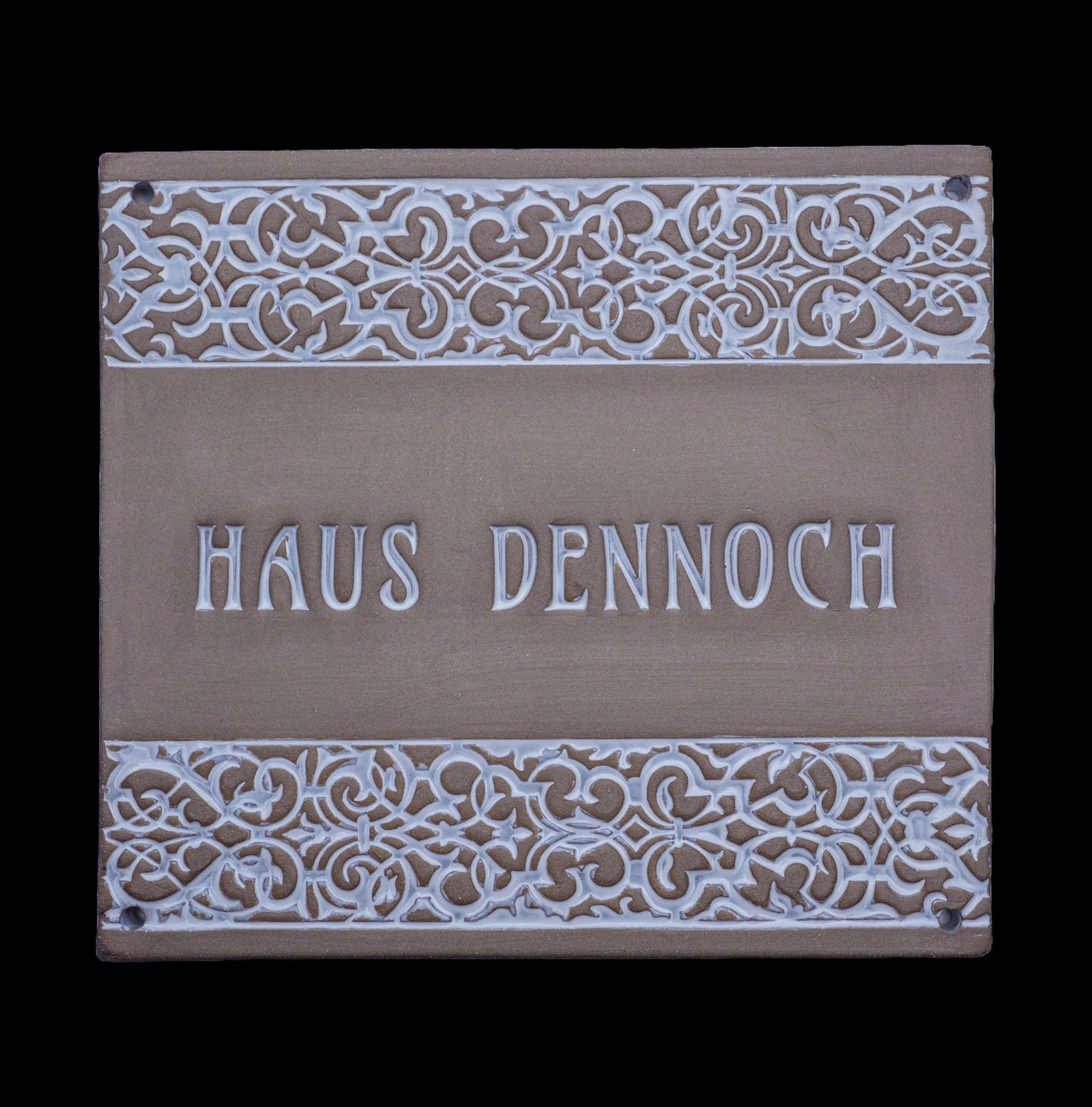 Rechteckiges dekoratives Hausschild mit verzierten floralen Mustern am oberen und unteren Rand und der Gravur „Haus Dennoch“ in der Mitte.