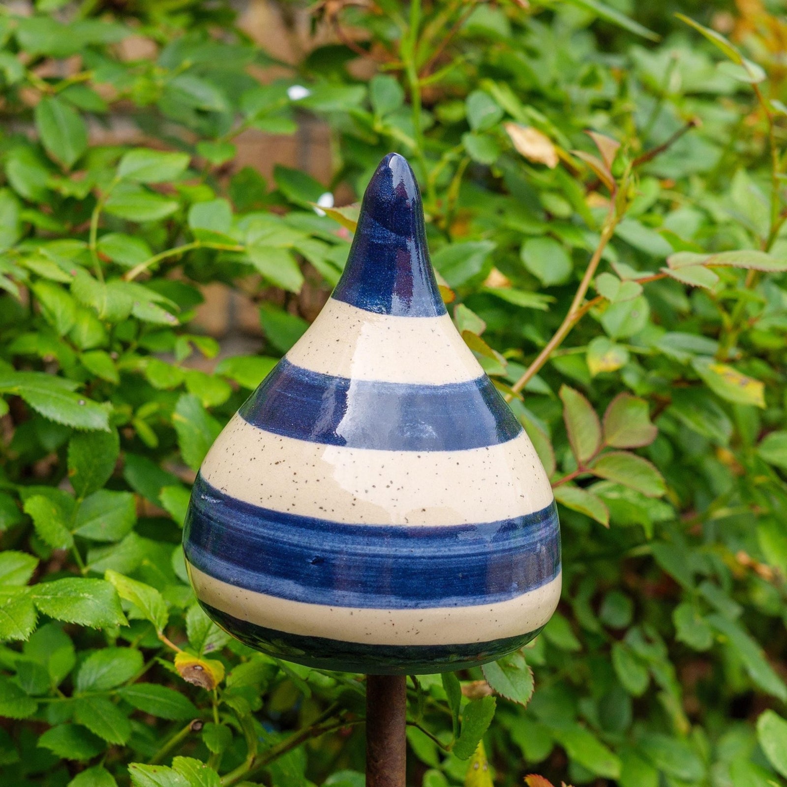 Handgefertigte Keramik-Glocke in Tropfenform mit glänzenden blauen und beigefarbenen Streifen, auf einem Holzstab befestigt, vor blühenden rosa Blumen.