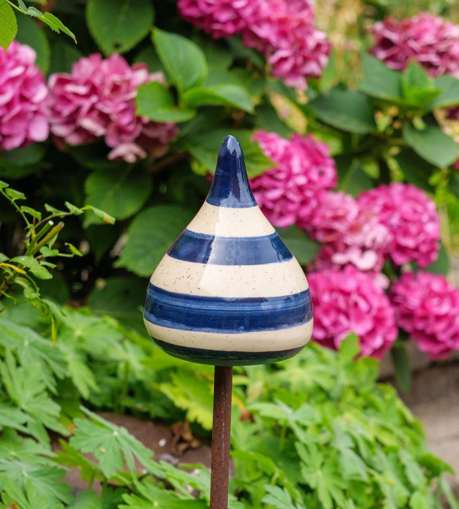 Handgefertigte Keramik-Glocke in Tropfenform mit glänzenden blauen und beigefarbenen Streifen, auf einem Holzstab befestigt, vor blühenden rosa Blumen.