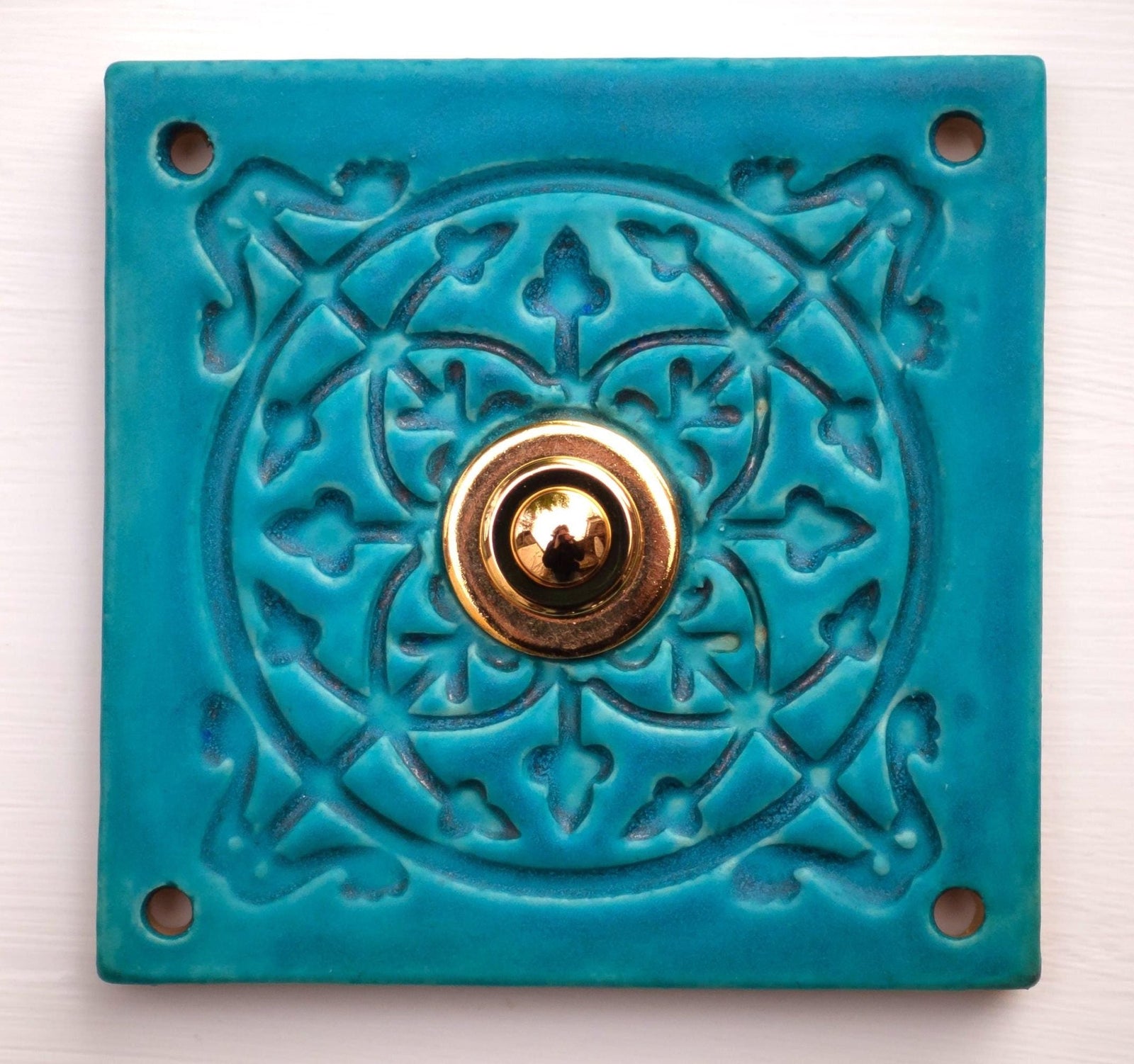 Quadratische Keramikklingel in Türkisblau mit geprägtem Mandala-Muster und goldenem Druckknopf in der Mitte