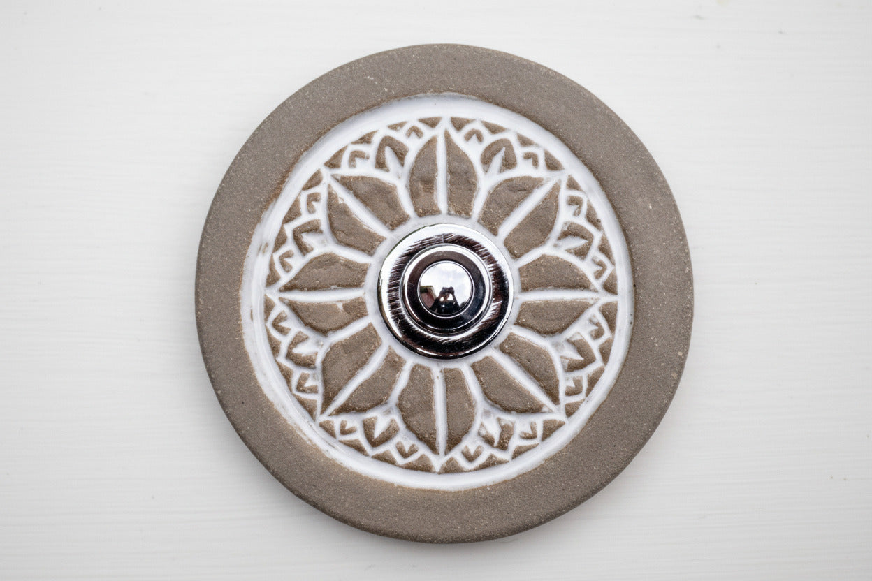 Runde dekorative Klingel mit geprägtem weißem Mandala-Blumenmuster auf graubrauner Platte und silbernem Druckknopf in der Mitte.
