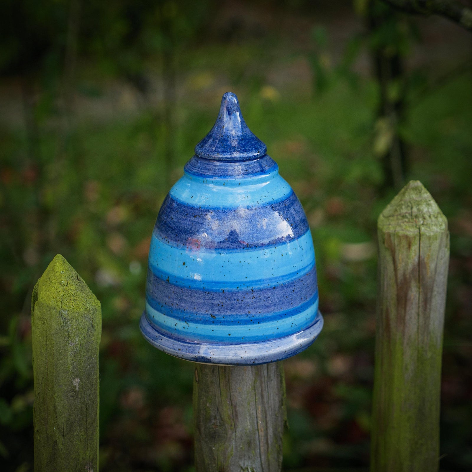 Handgefertigter blau-gestreifter Keramik-Pfostenhocker im Garten, glasiert in mehreren Blautönen mit horizontalen Streifen. Kegelförmige Gartenspitze, ca. 12 cm hoch, mit einer 5–6 cm großen Öffnung unten – passend für Zaunpfähle oder Holzstäbe. Das Stück steht auf einem Gartenzaun zwischen grünen Pflanzen im Freien.