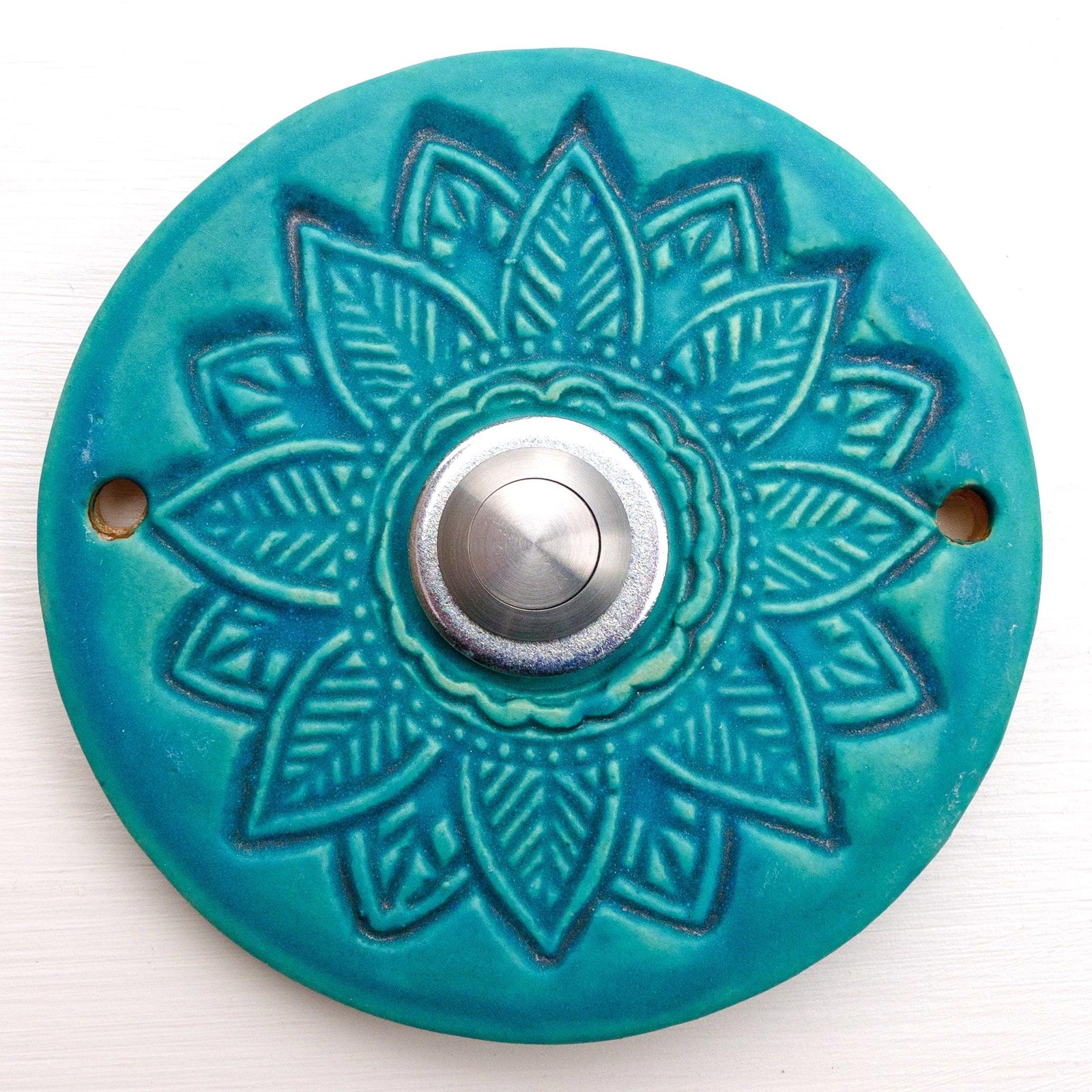 Runde dekorative Klingel mit geprägtem Mandala-Blumenmuster auf blauer Platte und silbernem Druckknopf in der Mitte.