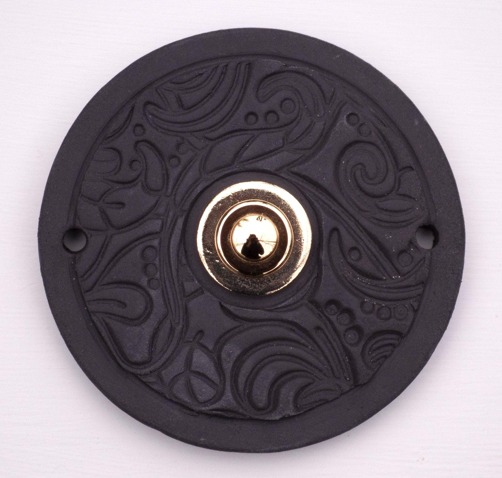 Runde dekorative Klingel mit dezentem geprägtem Wirbelmuster auf matter schwarzer Platte und goldenem Druckknopf in der Mitte.
