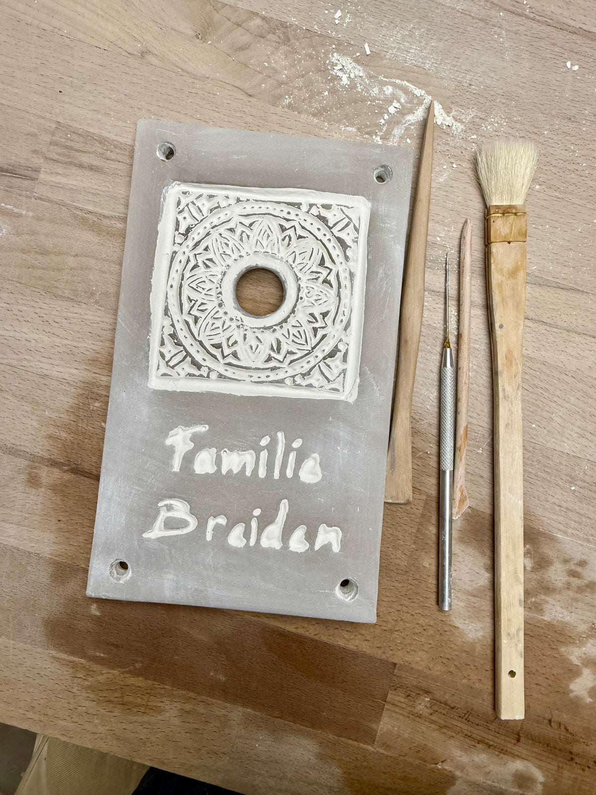 a wooden table with various crafting tools and materials, including a white plaque with a decorative design, a paintbrush, and a knife.