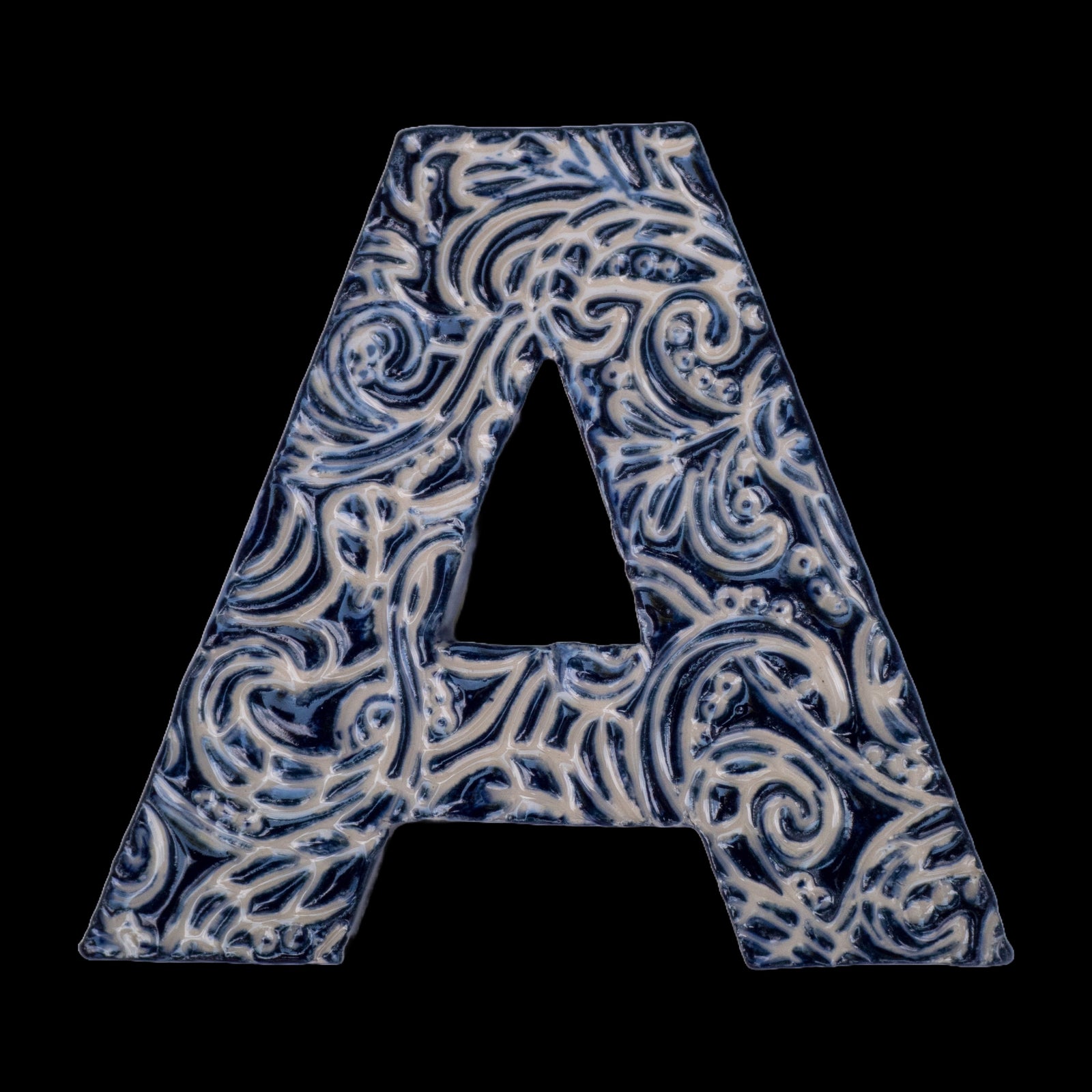 Handgefertigter Keramikbuchstabe „A“ in Dunkelblau mit beigefarbenen Ornamentmustern, vor schwarzem Hintergrund dargestellt.