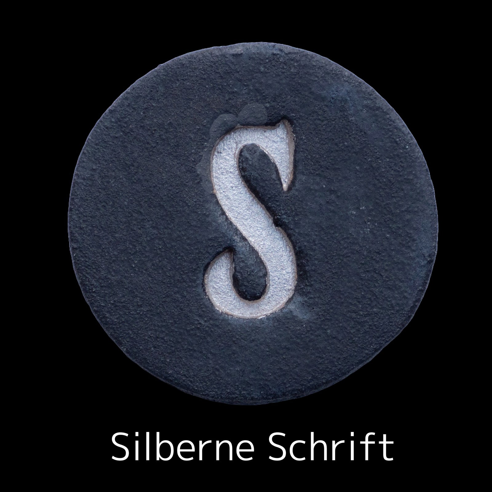 Runde dunkle Keramikplatte mit graviertem silbernen Buchstaben S in der Mitte, beschriftet mit 'Silberne Schrift