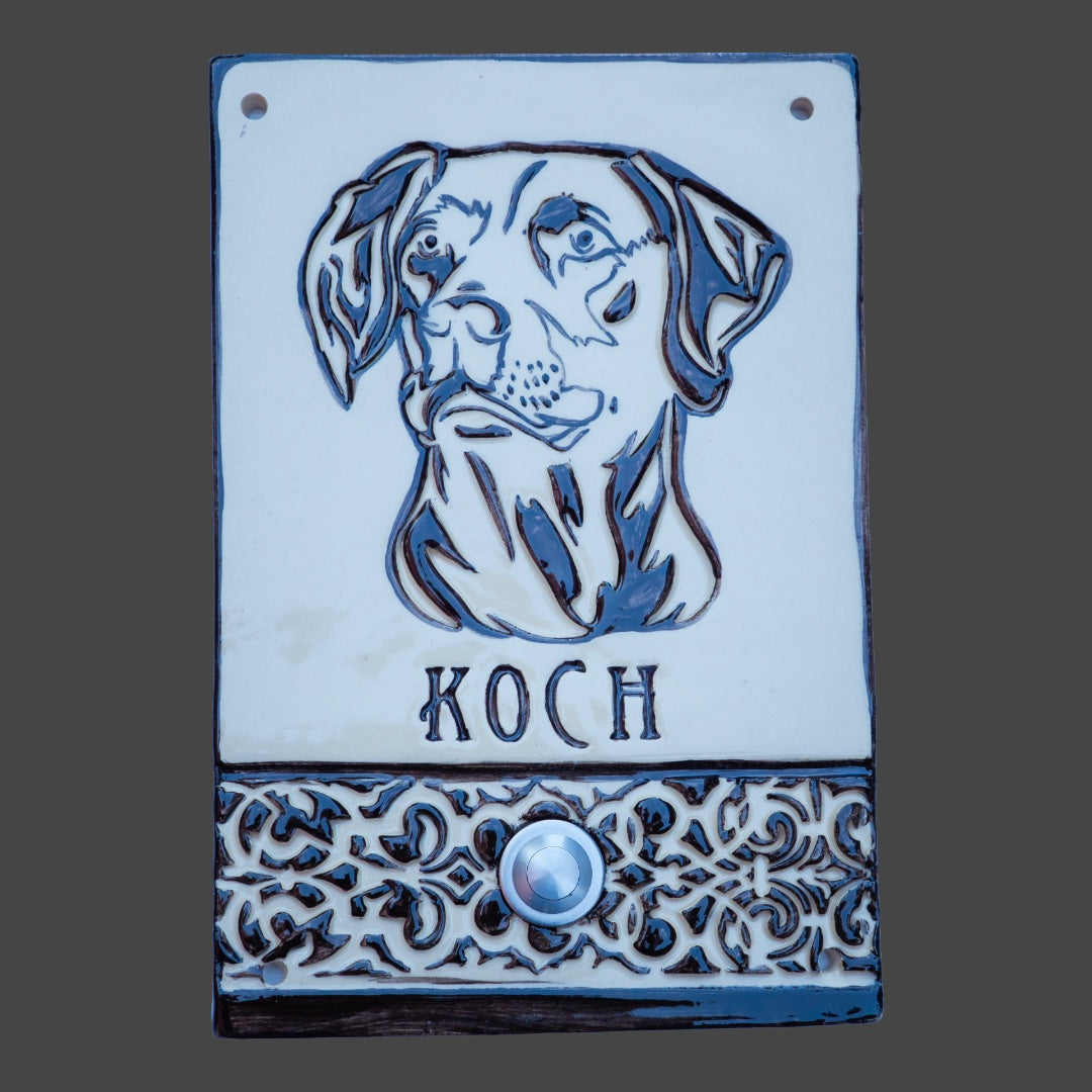Rechteckige Keramiktürklingelplatte mit schwarzer Hundegravur über dem Namen 'Koch'. Der untere Bereich zeigt ein dekoratives Muster mit einem silbernen Knopf in der Mitte