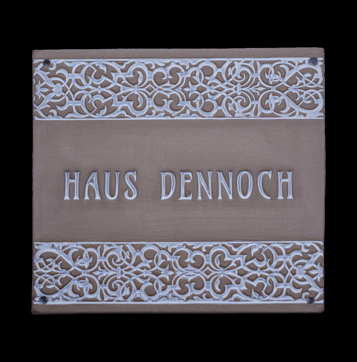 Rechteckiges dekoratives Hausschild mit verzierten floralen Mustern am oberen und unteren Rand und der Gravur „Haus Dennoch“ in der Mitte.