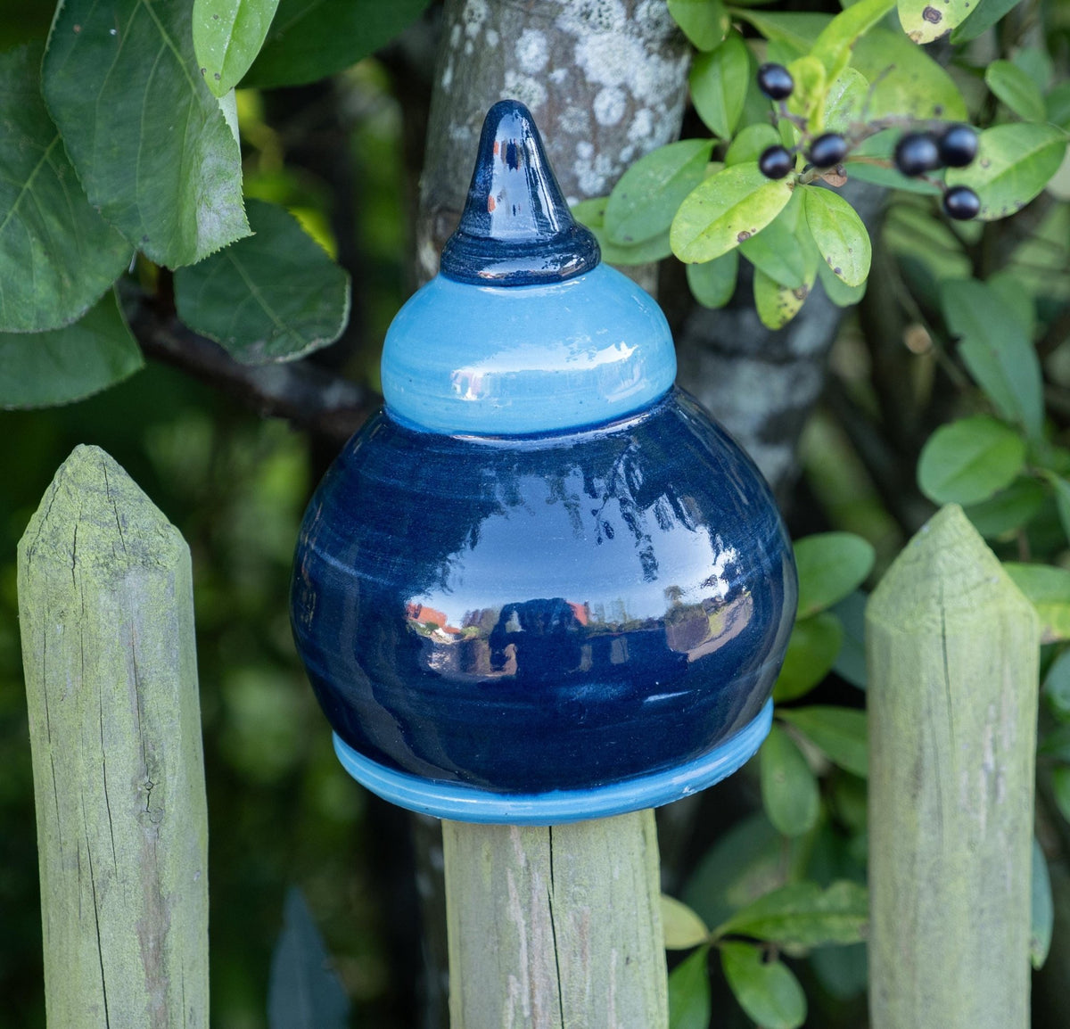 Handgefertigte Keramik-Glocke in glänzendem Dunkelblau mit hellblauem Abschluss, auf einem Holzpfosten vor einem Baum mit grünen Blättern und Beeren befestigt.