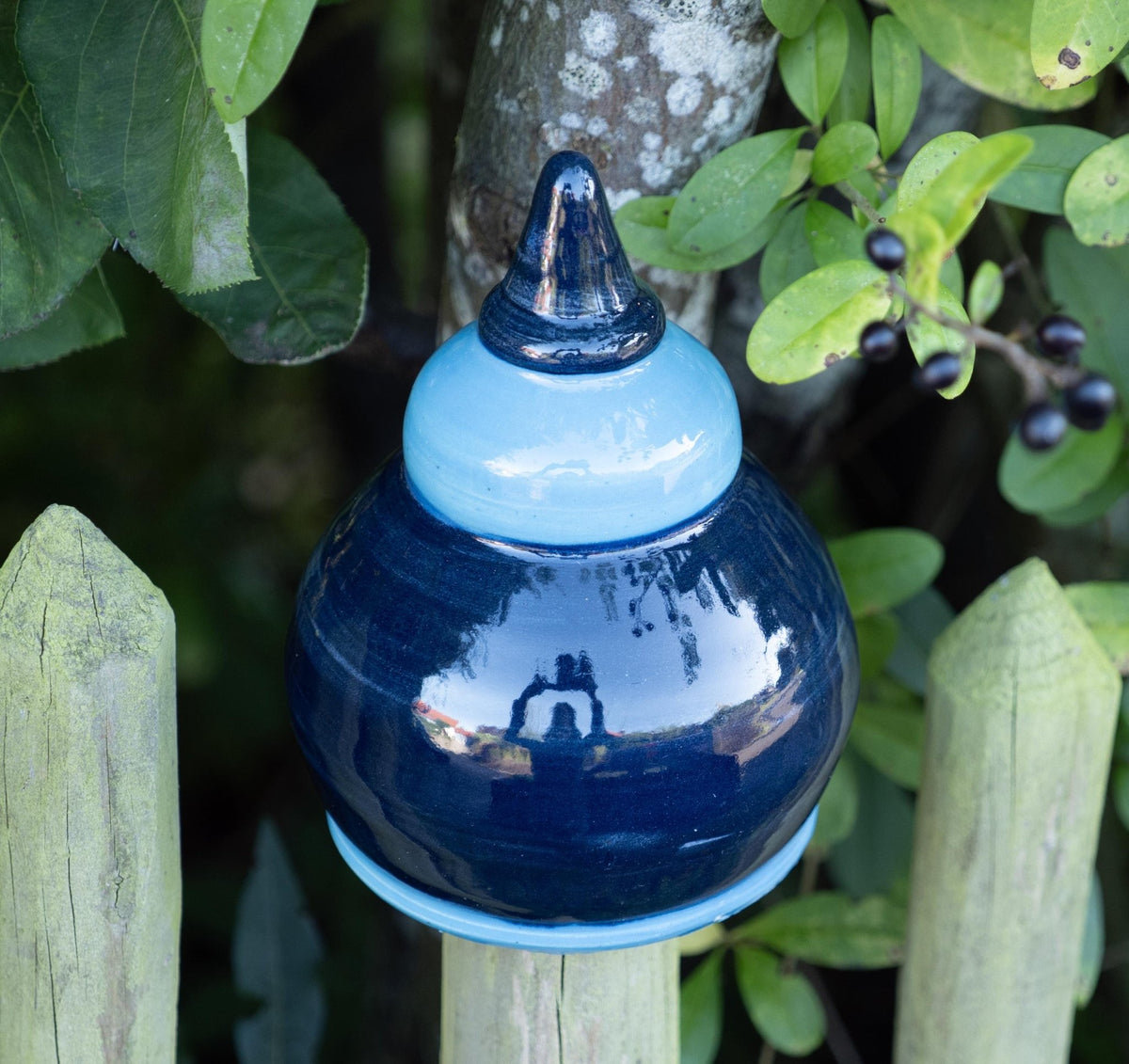 Handgefertigte Keramik-Glocke in glänzendem Dunkelblau mit hellblauem Abschluss, auf einem Holzpfosten vor einem Baum mit grünen Blättern und Beeren befestigt.