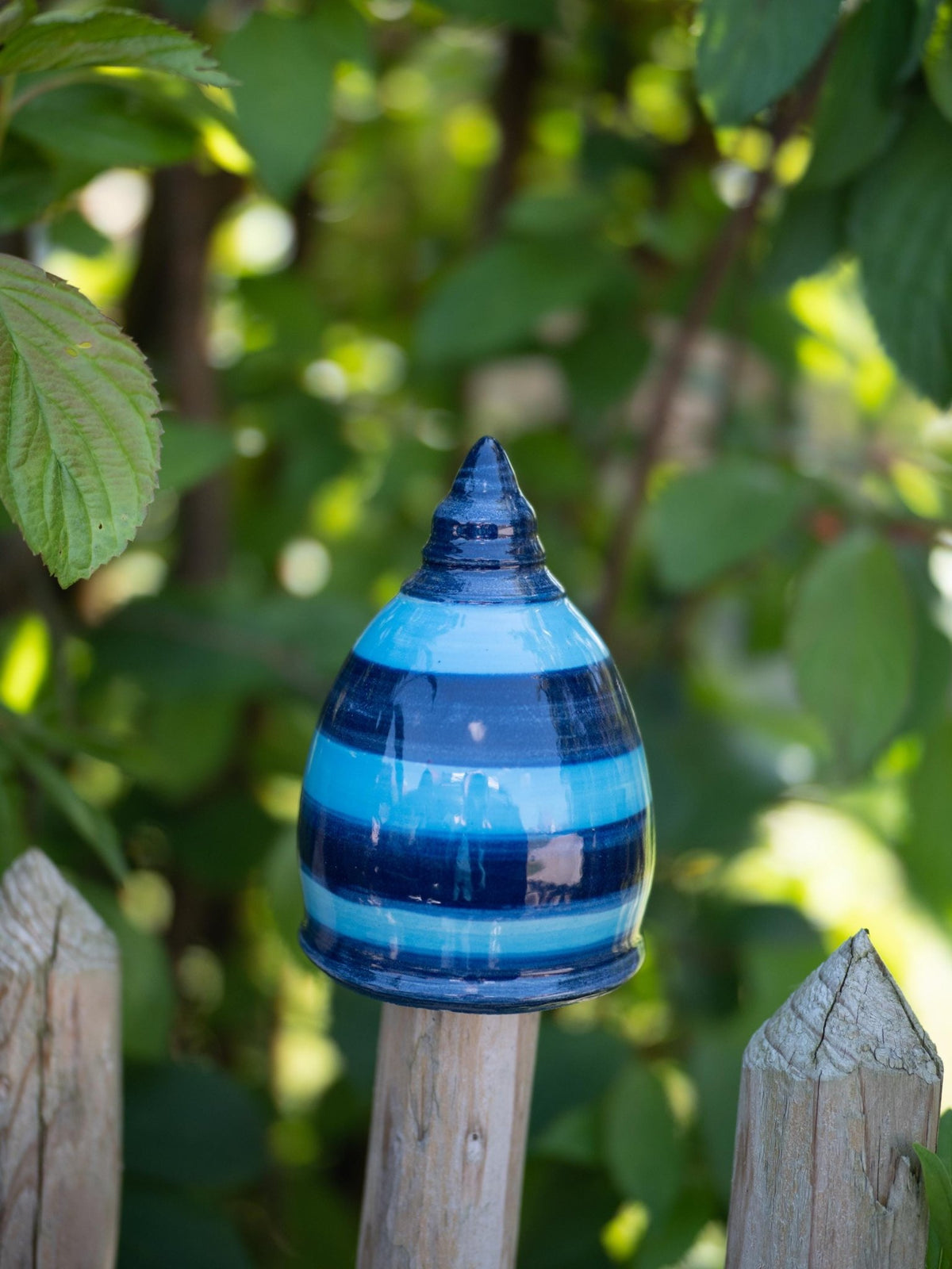 Handgefertigtes Keramik-Gartenornament in hoher Kegelform mit glänzender blau-hellblauer Streifenglasur, auf einem Holzzaunpfosten im Freien befestigt.