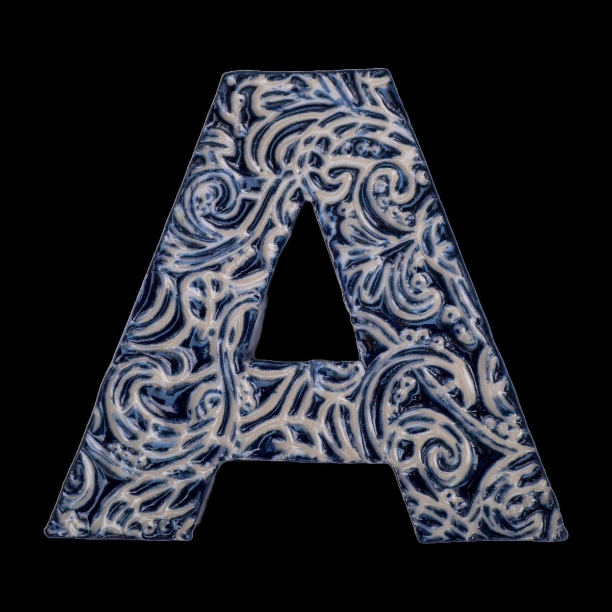 Handgefertigter Keramikbuchstabe „A“ in Dunkelblau mit beigefarbenen Ornamentmustern, vor schwarzem Hintergrund dargestellt.