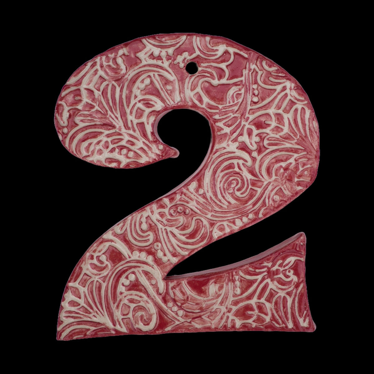 Handgefertigte Keramikzahl „2“ in Rot mit beigefarbenen Ornamentmustern, vor schwarzem Hintergrund dargestellt.