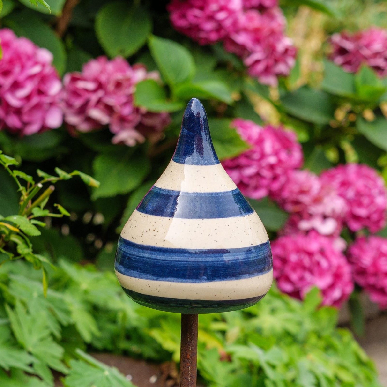 Handgefertigte Keramik-Glocke in Tropfenform mit glänzenden blauen und beigefarbenen Streifen, auf einem Holzstab befestigt, vor blühenden rosa Blumen.