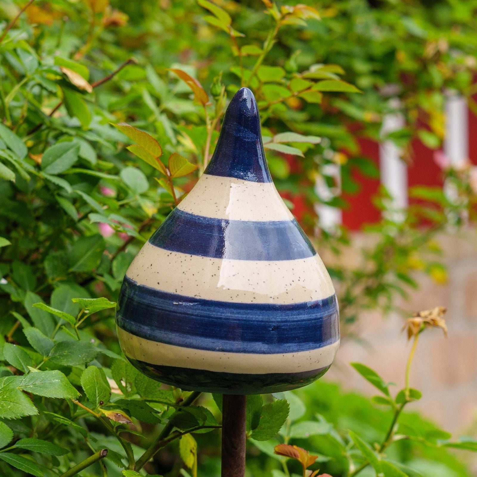 Handgefertigte Keramik-Glocke in Tropfenform mit glänzenden blauen und beigefarbenen Streifen, auf einem Holzstab befestigt, vor blühenden rosa Blumen.