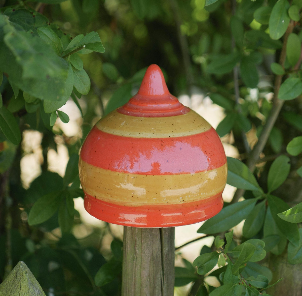 Handgefertigte Keramik-Glocke mit glänzenden orangefarbenen und gelben Streifen, auf einem Holzpfosten befestigt und von grünen Blättern umgeben.