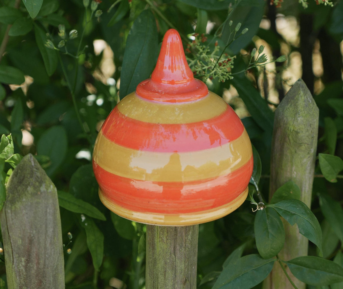 Handgefertigte Keramik-Glocke mit glänzenden orangefarbenen und gelben Streifen, auf einem Holzpfosten befestigt und von grünen Blättern umgeben.