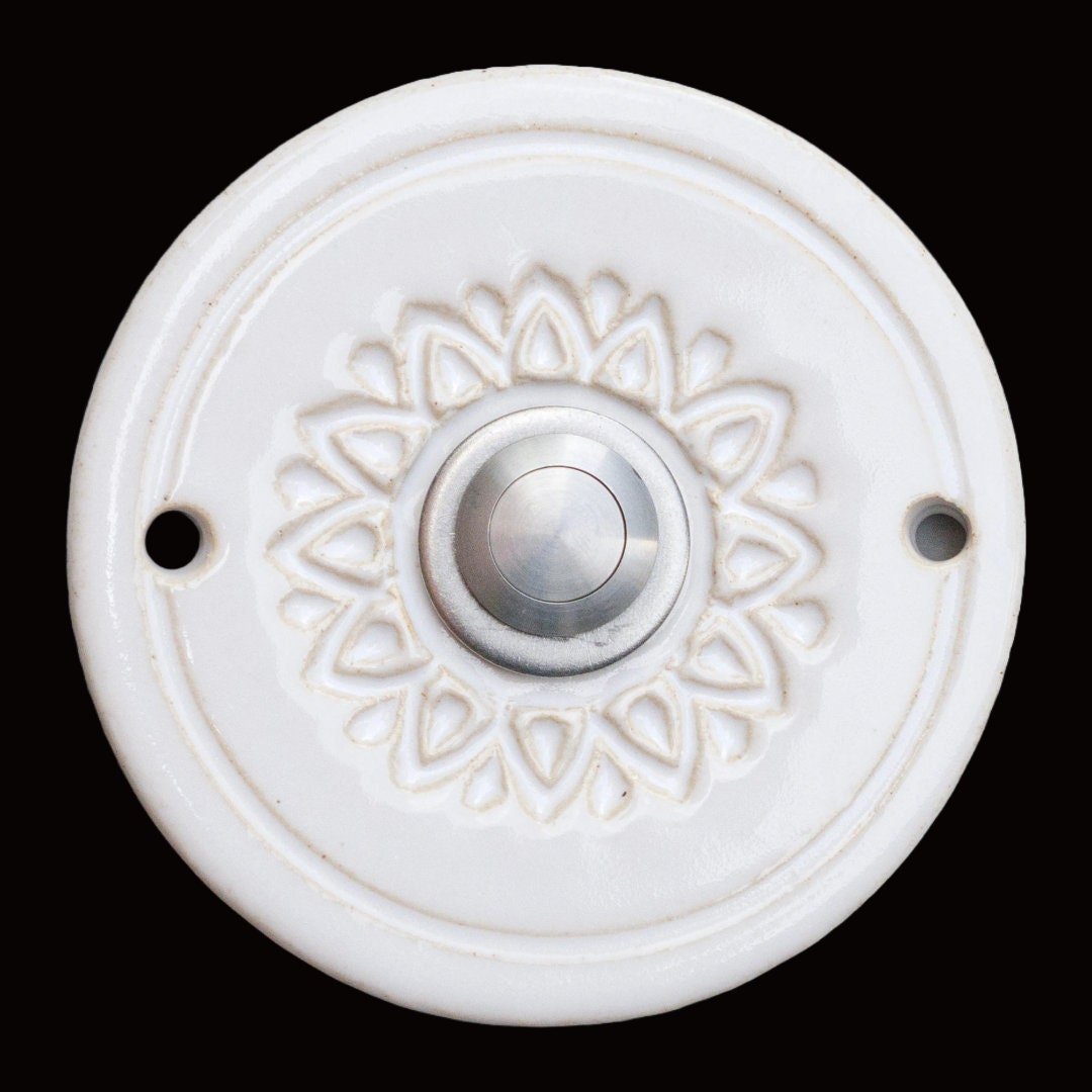 Runde dekorative Klingel mit geprägtem Blumen-Sternmuster auf weißer Platte und silbernem Druckknopf in der Mitte