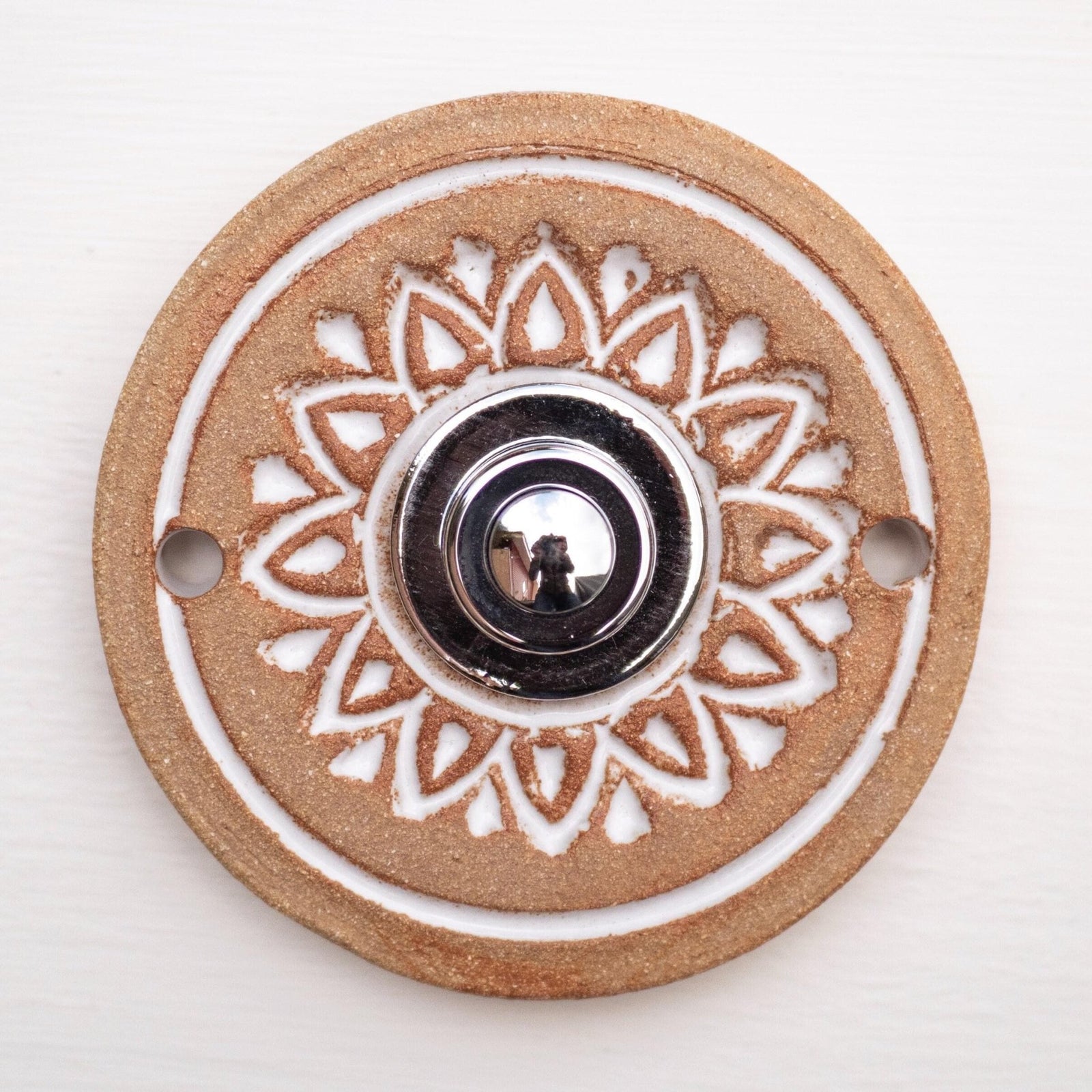 Runde dekorative Klingel mit weißem geprägtem Blumen-Sternmuster auf goldbrauner Platte und silbernem Druckknopf in der Mitte.