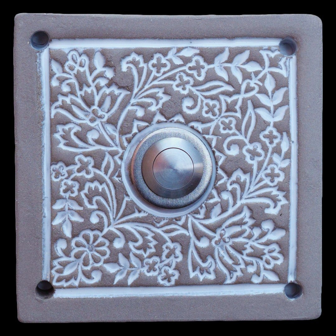Quadratische Keramik-Türklingel mit detaillierten floralen Mustern in weißem Relief und einem silbernen Knopf in der Mitte