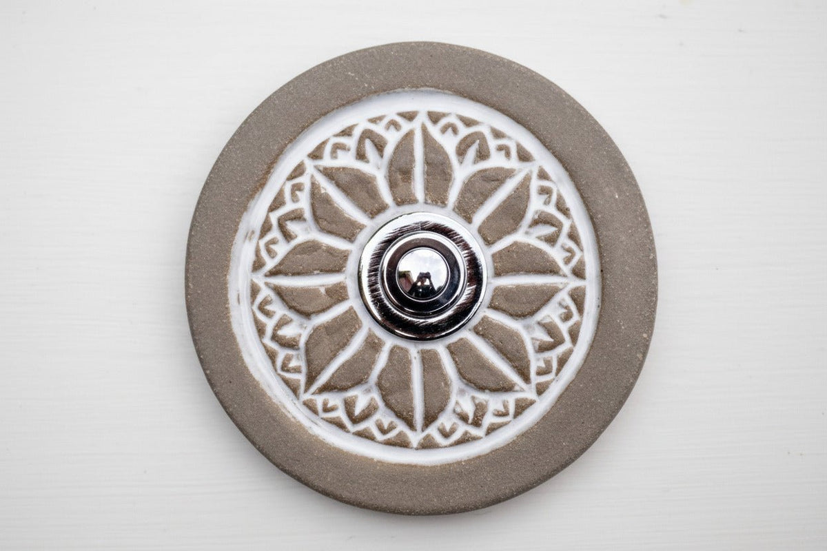 Runde dekorative Klingel mit geprägtem weißem Mandala-Blumenmuster auf graubrauner Platte und silbernem Druckknopf in der Mitte.