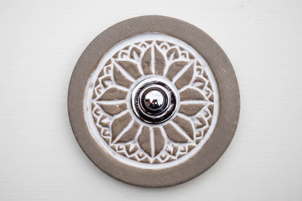 Runde dekorative Klingel mit geprägtem weißem Mandala-Blumenmuster auf graubrauner Platte und silbernem Druckknopf in der Mitte.