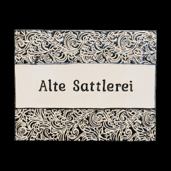 Handgefertigtes Keramikschild mit ornamentalem Schwarz-Weiß-Design und der Aufschrift „Alte Sattlerei“. Elegant und wetterfest für den Außenbereich.