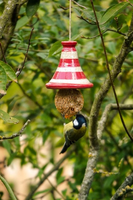 Rot-weiß gestreiftes Keramik-Vogelfutterhaus mit Meisenknödel, hängend im Freien vor grünem Waldhintergrund