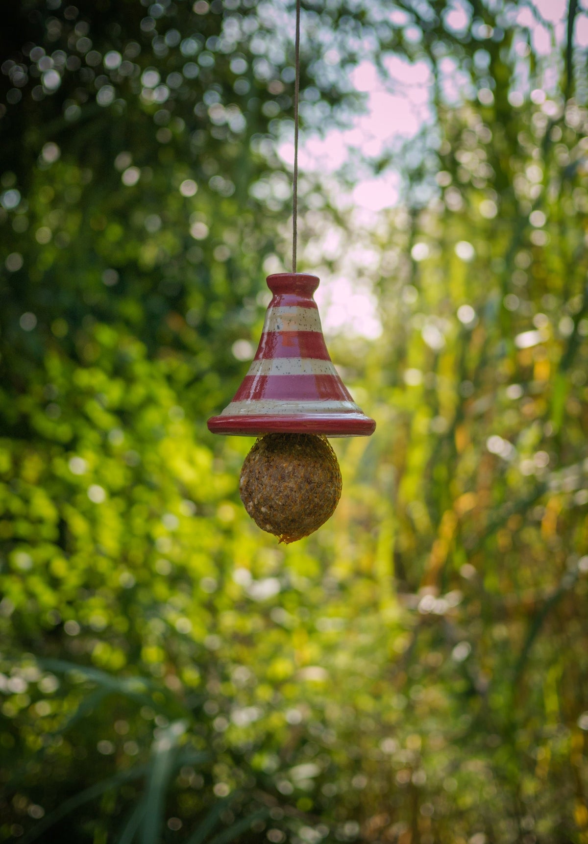 Rot-weiß gestreiftes Keramik-Vogelfutterhaus mit Meisenknödel, hängend im Freien vor grünem Waldhintergrund