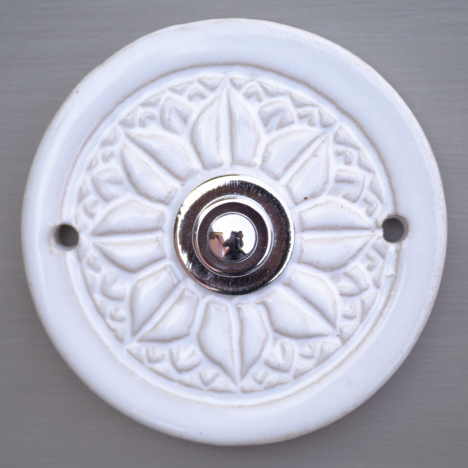 Runde dekorative Klingel mit geprägtem Mandala-Blumenmuster auf weißer Platte und silbernem Druckknopf in der Mitte