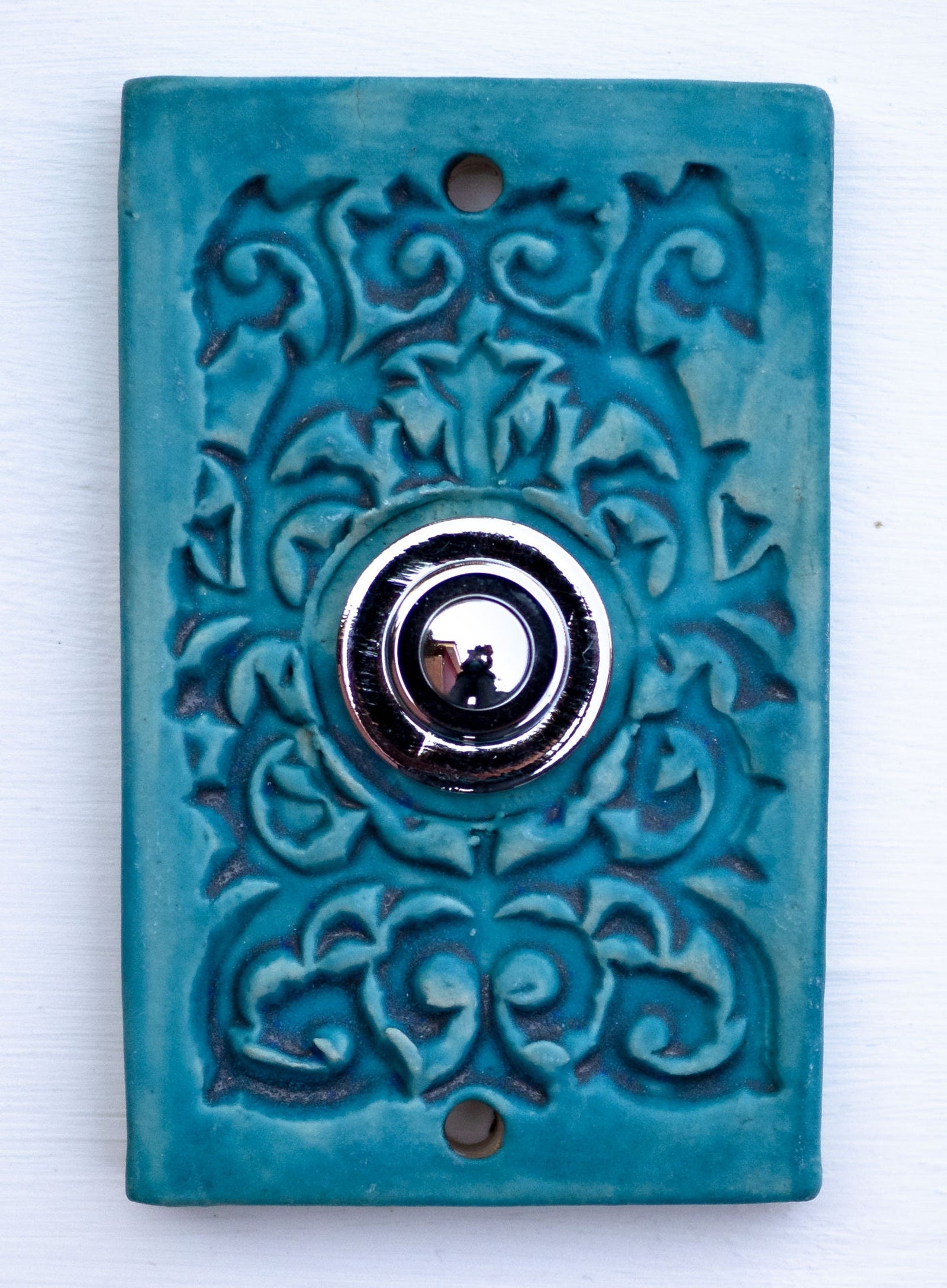Rechteckige Keramikklingel in Türkisblau mit kunstvollem Reliefmuster und zentralem silbernem Druckknopf