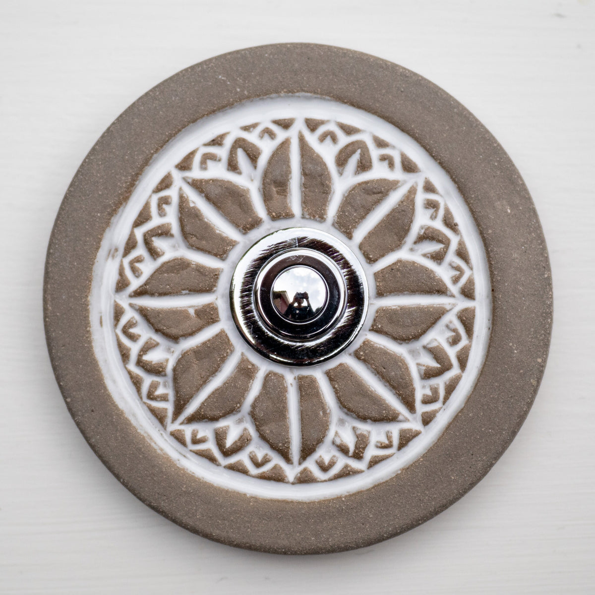 Runde dekorative Klingel mit geprägtem weißem Mandala-Blumenmuster auf graubrauner Platte und silbernem Druckknopf in der Mitte.