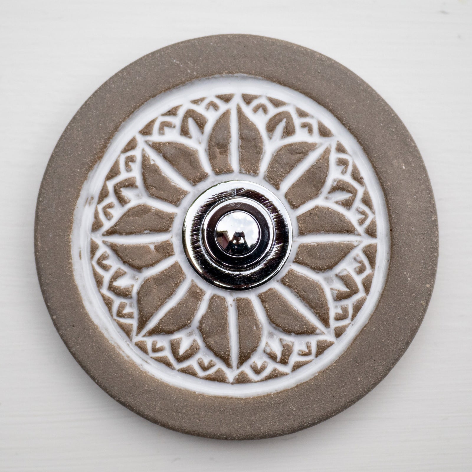 Runde dekorative Klingel mit geprägtem weißem Mandala-Blumenmuster auf graubrauner Platte und silbernem Druckknopf in der Mitte.