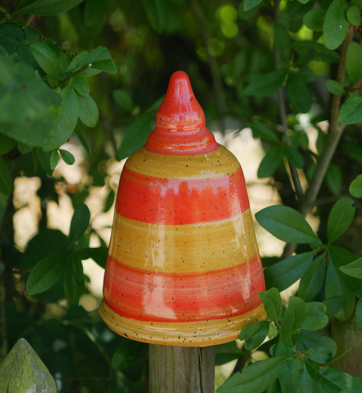Bunte Keramik-Gartendekoration in Glockenform mit orangefarbenen und gelben Streifen, zwischen grünen Pflanzen und Holzpfosten platziert.