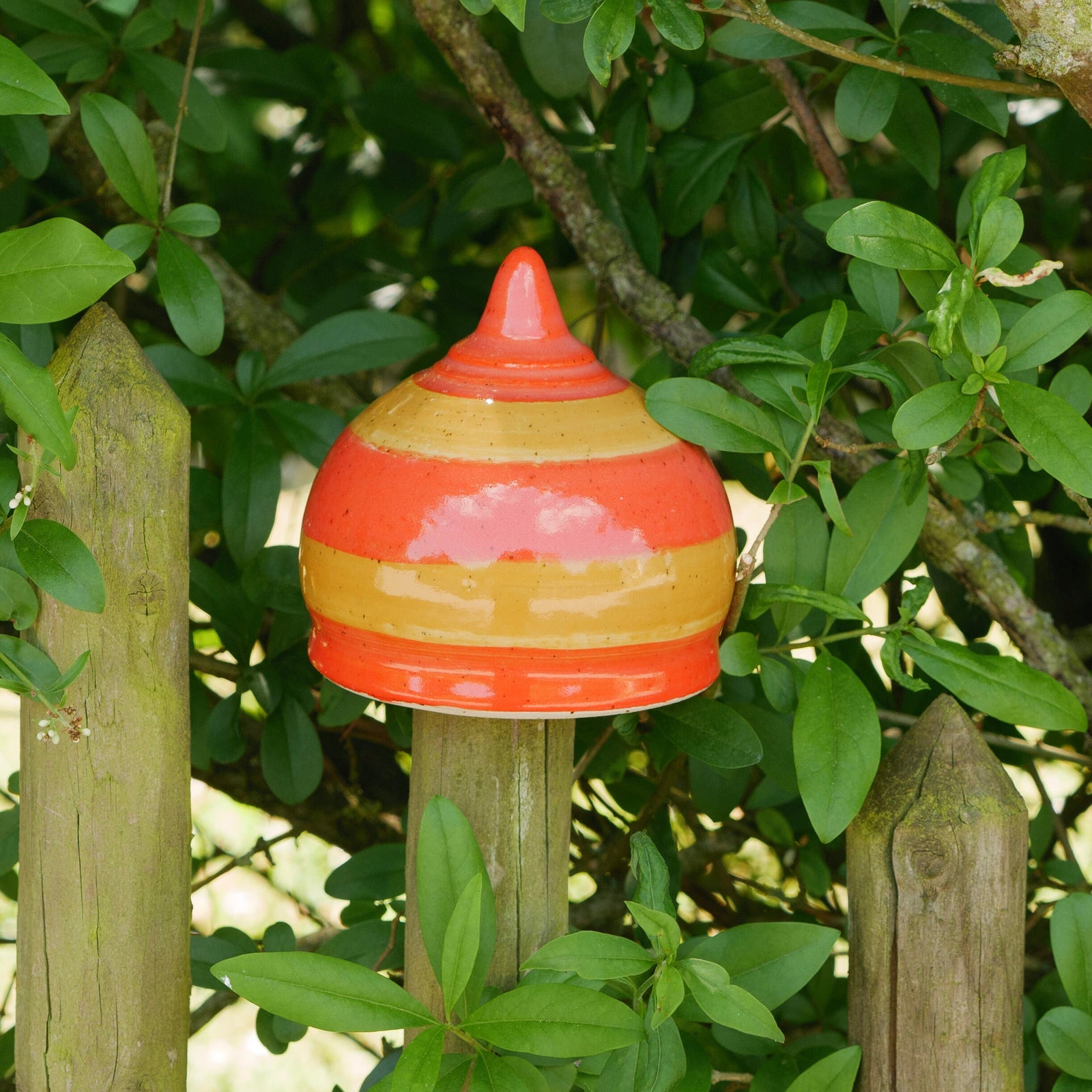 Bunte Keramik-Gartendekoration in Kegelform mit orangefarbenen und gelben Streifen, zwischen grünen Pflanzen und Holzpfosten platziert