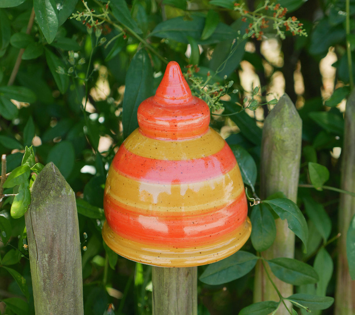 Handgefertigte Keramik-Glocke mit glänzenden orangefarbenen und gelben Streifen, befestigt auf einem Holzpfosten, umgeben von grünen Blättern.