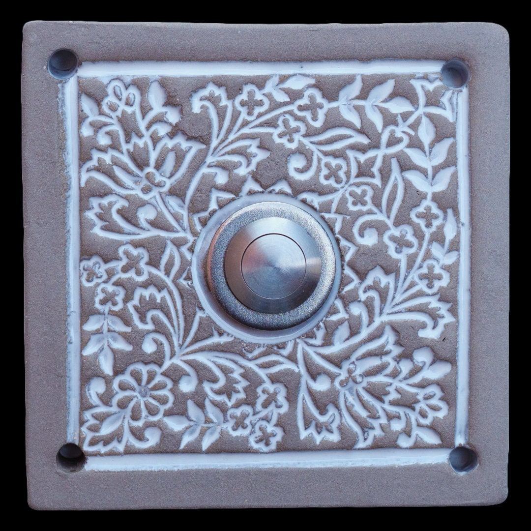 Quadratische Keramik-Türklingel mit detaillierten floralen Mustern in weißem Relief und einem silbernen Knopf in der Mitte