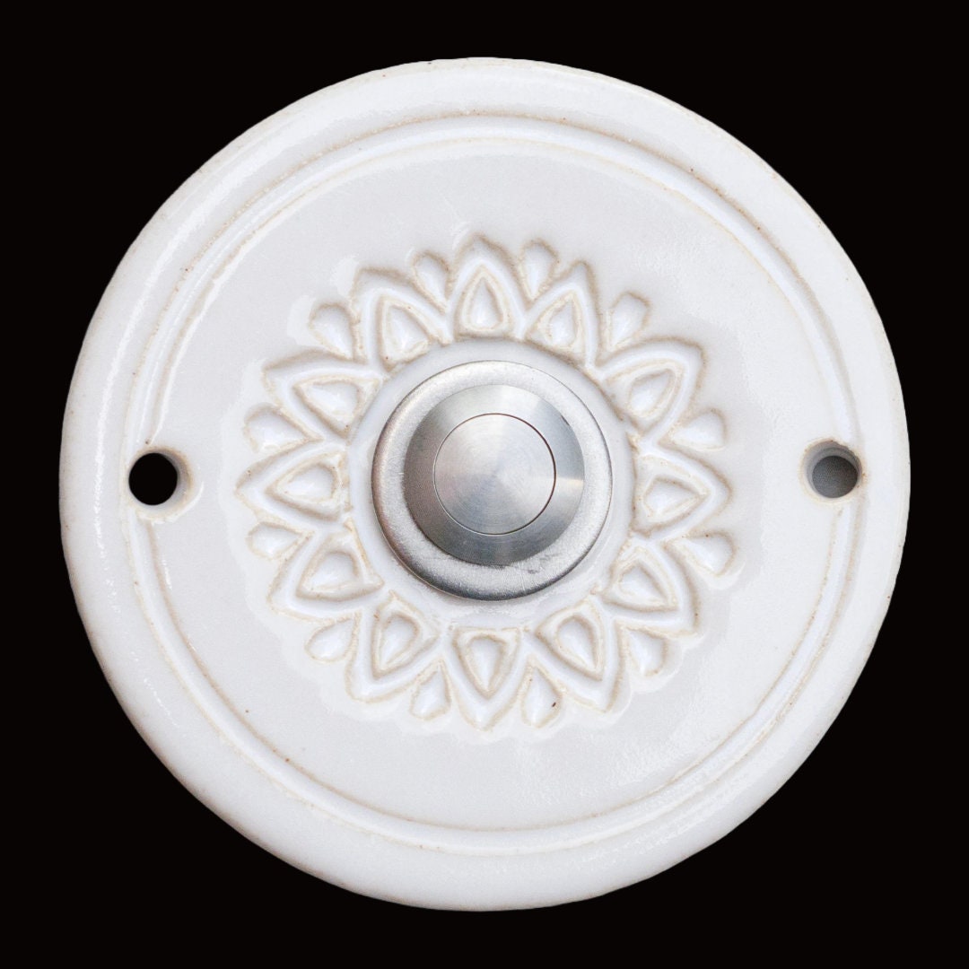 Runde dekorative Klingel mit geprägtem Blumen-Sternmuster auf weißer Platte und silbernem Druckknopf in der Mitte