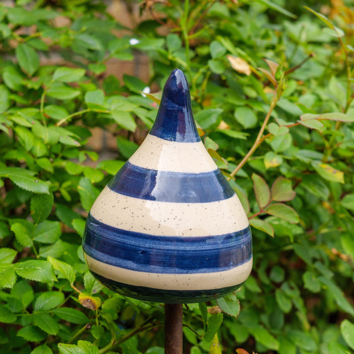 Handgefertigte Keramik-Glocke in Tropfenform mit glänzenden blauen und beigefarbenen Streifen, auf einem Holzstab befestigt, vor blühenden rosa Blumen.