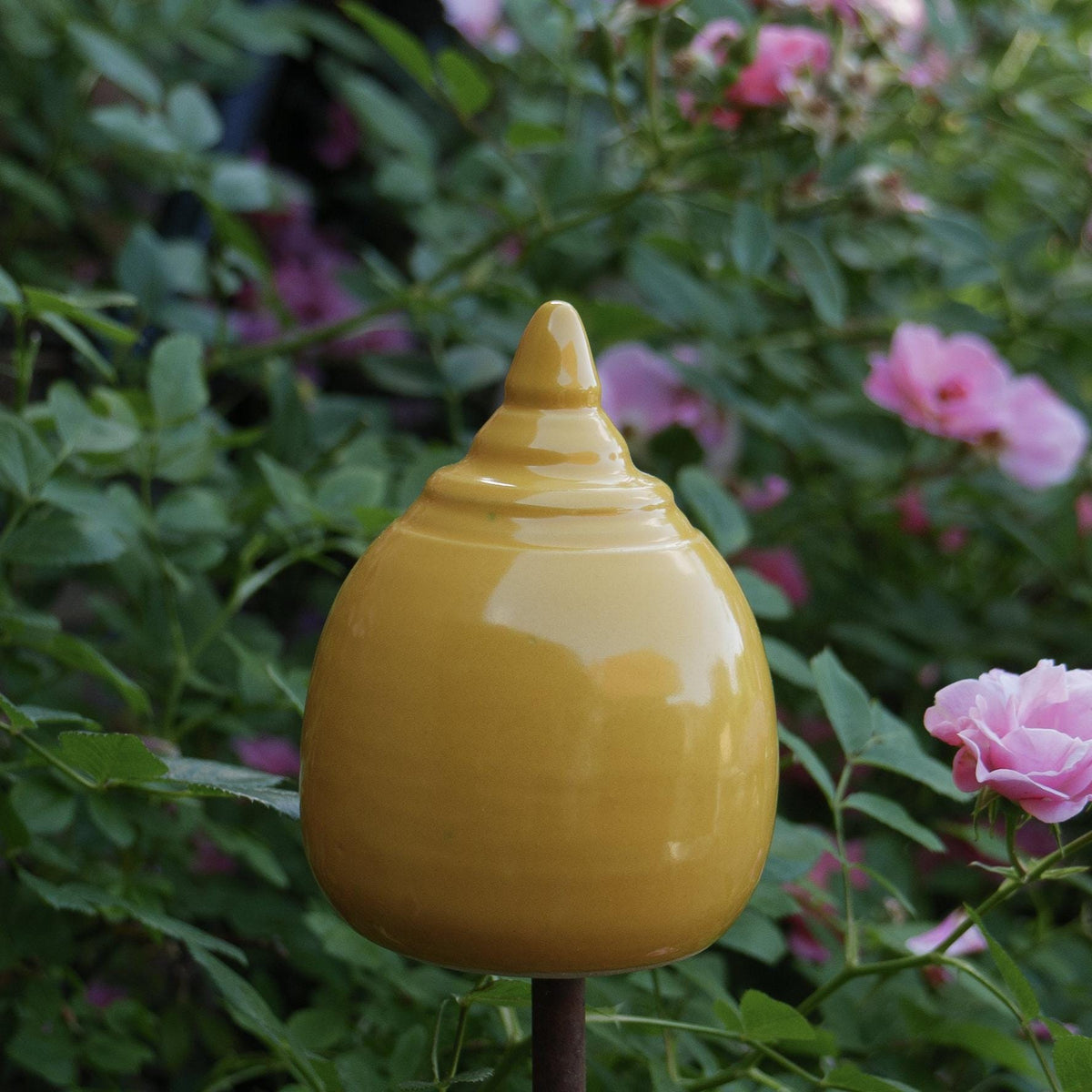 Handgefertigte Keramik-Gartenstele in glänzendem Senfgelb mit abgerundeter Kegelform, aufgestellt zwischen blühenden rosa Rosen