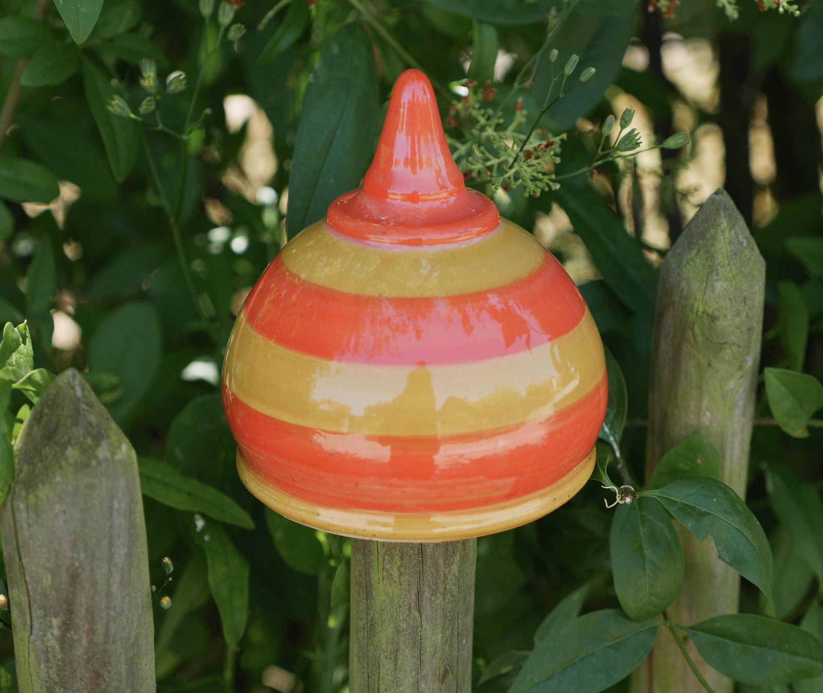 Handgefertigte Keramik-Glocke mit glänzenden orangefarbenen und gelben Streifen, auf einem Holzpfosten befestigt und von grünen Blättern umgeben.