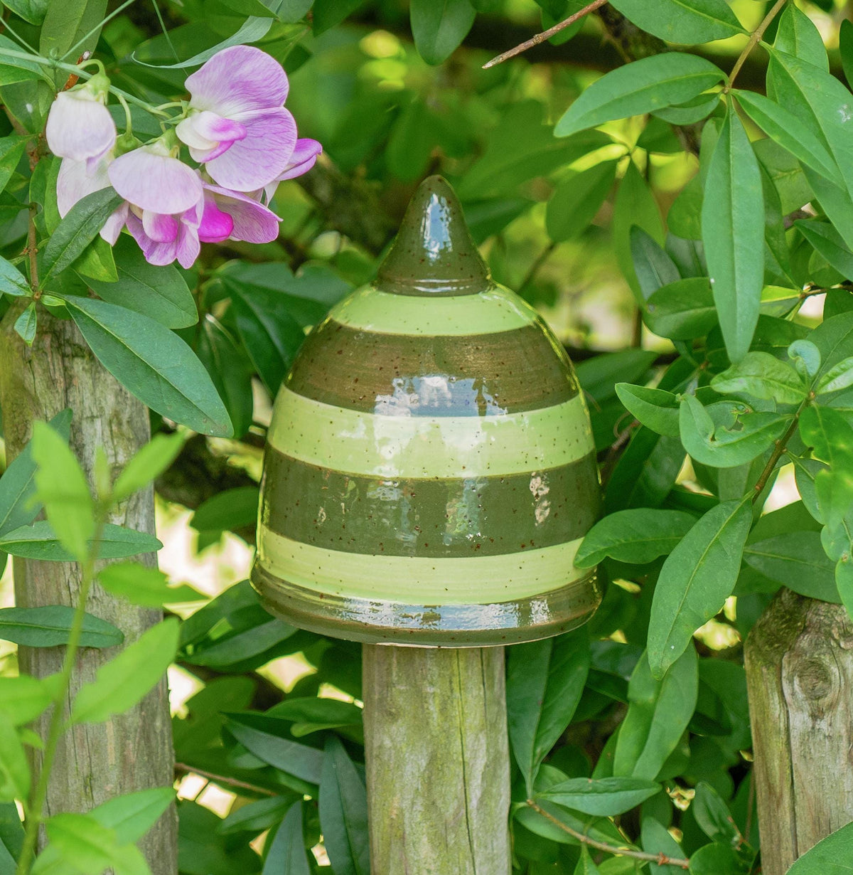 Handgefertigtes Keramik-Gartenornament in abgerundeter Kegelform mit glänzender grün-brauner Streifenglasur, auf einem Holzzaun zwischen Blättern befestigt.