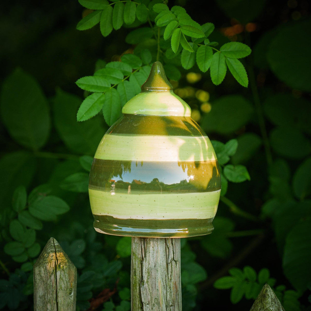 Handgefertigtes Keramik-Gartenornament in abgerundeter Kegelform mit glänzender grün-brauner Streifenglasur, auf einem Holzzaun zwischen Blättern befestigt.
