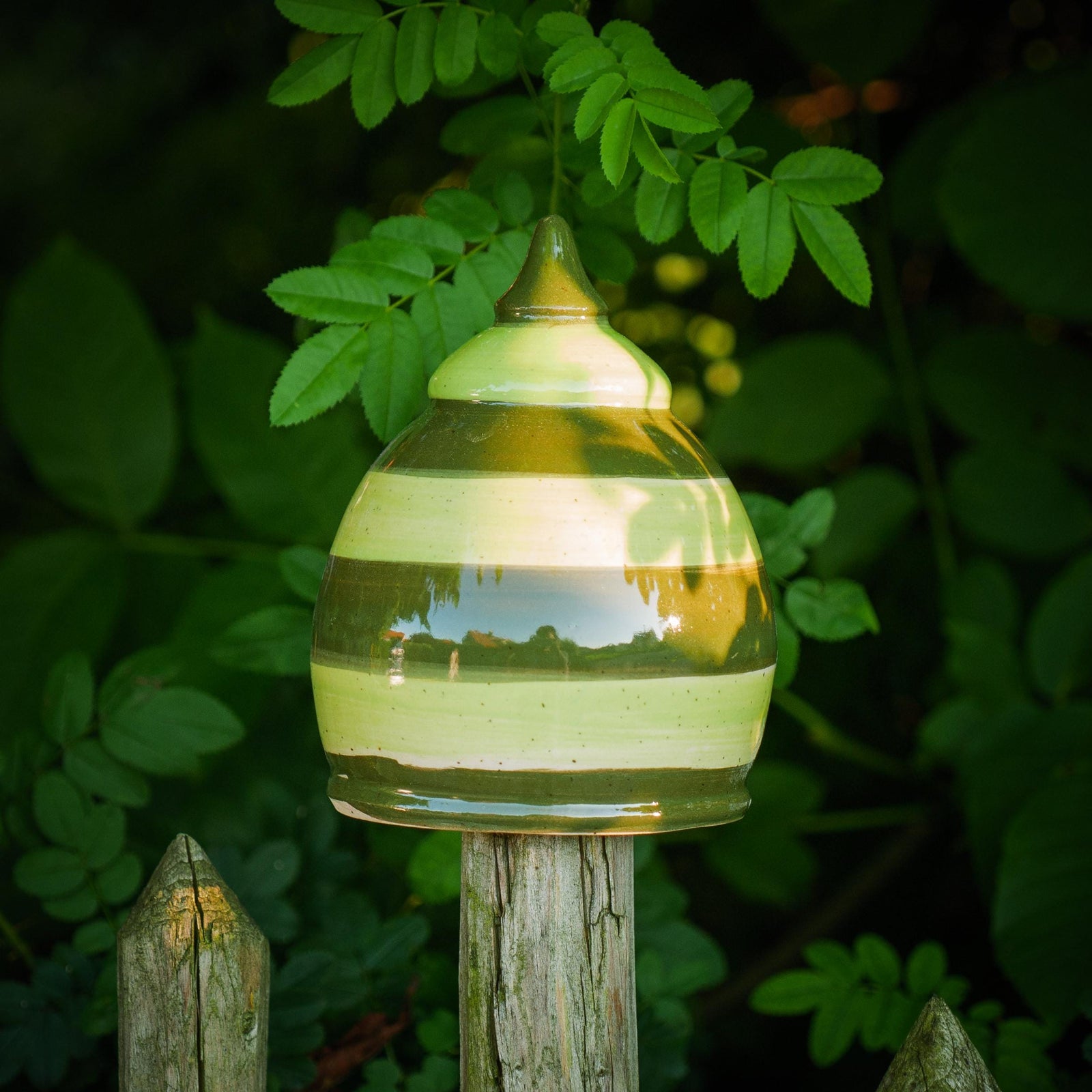 Handgefertigtes Keramik-Gartenornament in abgerundeter Kegelform mit glänzender grün-brauner Streifenglasur, auf einem Holzzaun zwischen Blättern befestigt.