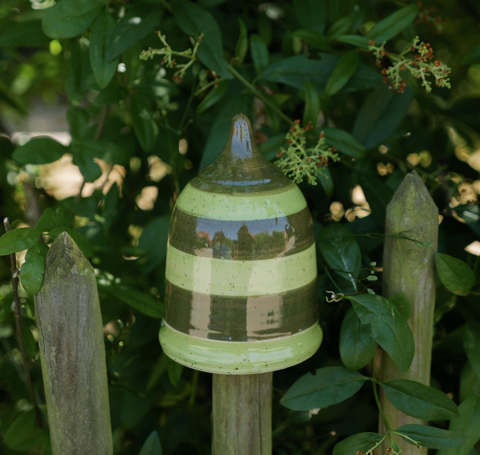 Handgefertigtes Keramik-Gartenornament in abgerundeter Kegelform mit glänzender grün-brauner Streifenglasur, auf einem Holzzaun zwischen Blättern befestigt.