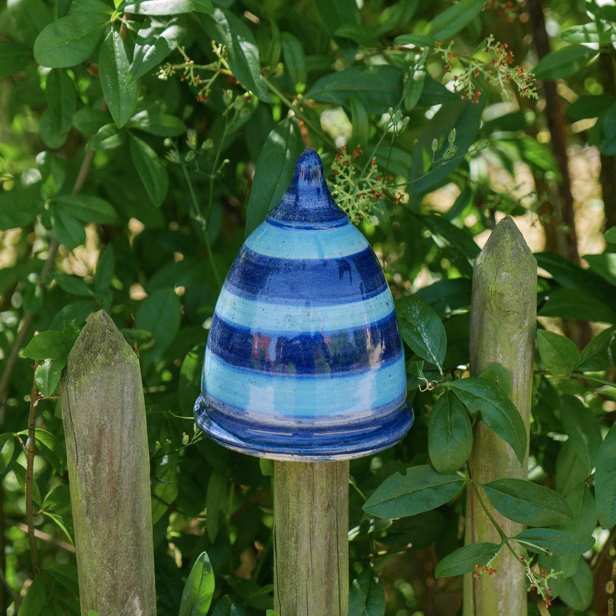 Handgefertigter blau-gestreifter Keramik-Pfostenhocker im Garten, glasiert in mehreren Blautönen mit horizontalen Streifen. Kegelförmige Gartenspitze, ca. 12 cm hoch, mit einer 5–6 cm großen Öffnung unten – passend für Zaunpfähle oder Holzstäbe. Das Stück steht auf einem Gartenzaun zwischen grünen Pflanzen im Freien.